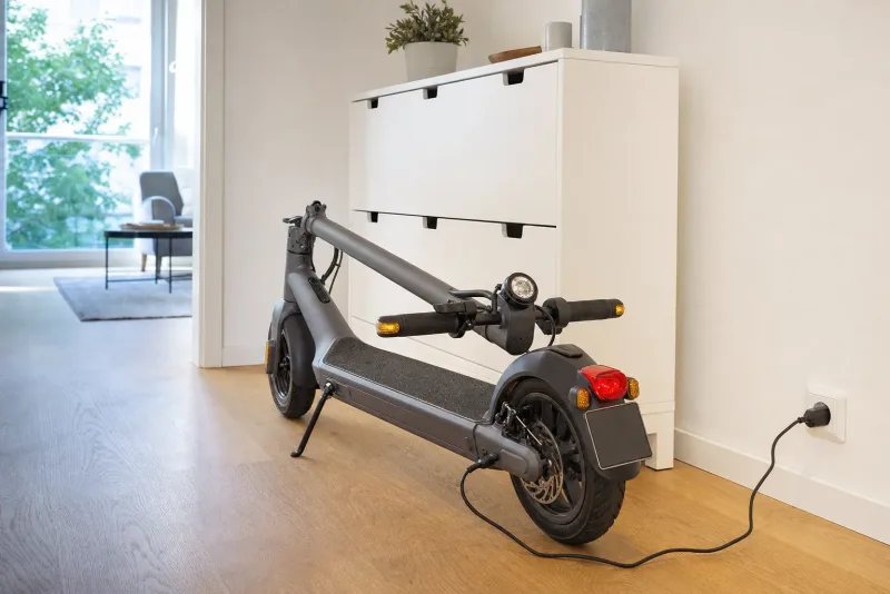 Zusammengeklappter E-Scooter mit schwarzer Versicherungsplakette am Heck in einem hellen Hausflur neben einer Steckdose