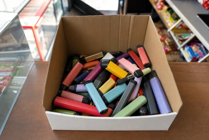 Zahlreiche bunte, ausgediente Einweg-E-Zigaretten gesammelt in einer Pappbox auf einer Ladentheke