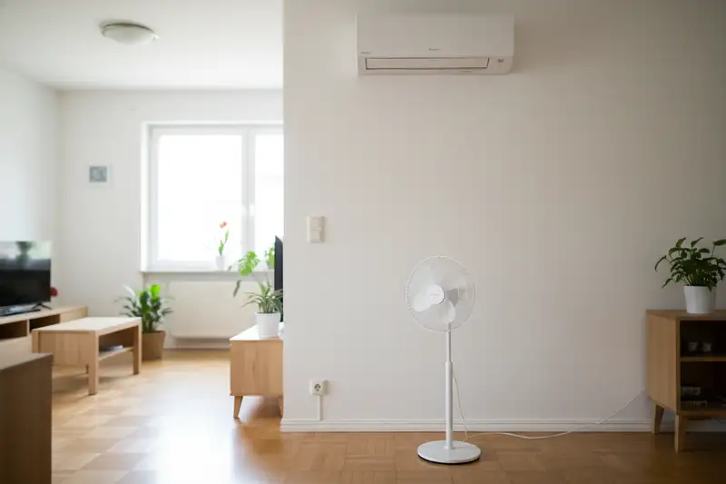 Wandmontierte Split-Klimaanlage über einem weißen Standventilator in einem hellen Wohnzimmer mit Holzboden