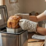 Brotbackautomaten im Praxis-Check: Diese 8 Fehler sollten Sie vermeiden Eine Frau mit Ofenhandschuhen nimmt ein frisch gebackenes Brot aus einem Brotbackautomaten, Dampf steigt auf. Sie lächelt.