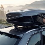 Eine Person verstaut eine Reisetasche in der geöffneten Thule-Dachbox auf einem Auto vor nebligen Bergen.