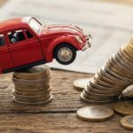 Roter Spielzeug-Käfer steht auf einem Stapel Euromünzen, daneben ein umfallender Münzturm auf Holzgrund.