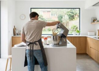 Ein Mann mit Schürze bedient eine Küchenmaschine auf einer Kochinsel in einer hellen Küche.