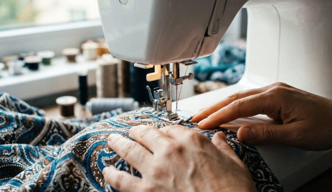 Eine Person näht mit einer Nähmaschine auf einem Stoff mit blauem und braunem Muster. Die Hände führen den Stoff unter der Nadel. Im Hintergrund sind Garnrollen zu sehen.