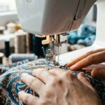 Die beste Nähmaschine - Test, Marktanalyse & Vergleich 12/2025 Eine Person näht mit einer Nähmaschine auf einem Stoff mit blauem und braunem Muster. Die Hände führen den Stoff unter der Nadel. Im Hintergrund sind Garnrollen zu sehen.