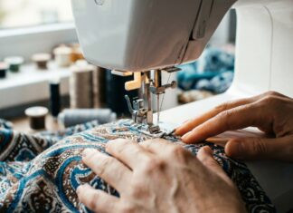 Eine Person näht mit einer Nähmaschine auf einem Stoff mit blauem und braunem Muster. Die Hände führen den Stoff unter der Nadel. Im Hintergrund sind Garnrollen zu sehen.