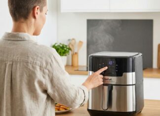 Eine Person bedient eine Heißluftfritteuse in einer Küche. Die Fritteuse ist eingeschaltet und Dampf steigt auf. Auf dem Tisch liegen Gemüse.