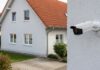 Weiße Überwachungskamera an einer Hauswand, im Hintergrund ein unscharfes Wohnhaus mit rotem Dach.