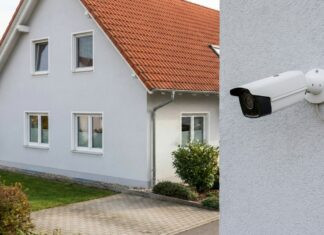 Weiße Überwachungskamera an einer Hauswand, im Hintergrund ein unscharfes Wohnhaus mit rotem Dach.