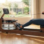 Das beste Wasserrudergerät aus Holz – Test, Marktanalyse & Vergleich 11/2025 Eine Frau trainiert auf einem hölzernen Rudergerät mit Wassertank in einem hellen Wohnzimmer.